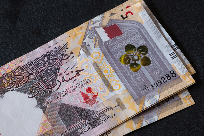 Close-up of two Qatari five-riyal banknotes, featuring intricate Arabic calligraphy, geometric patterns, and national symbols, including the Qatari flag and crest, set against a dark background.