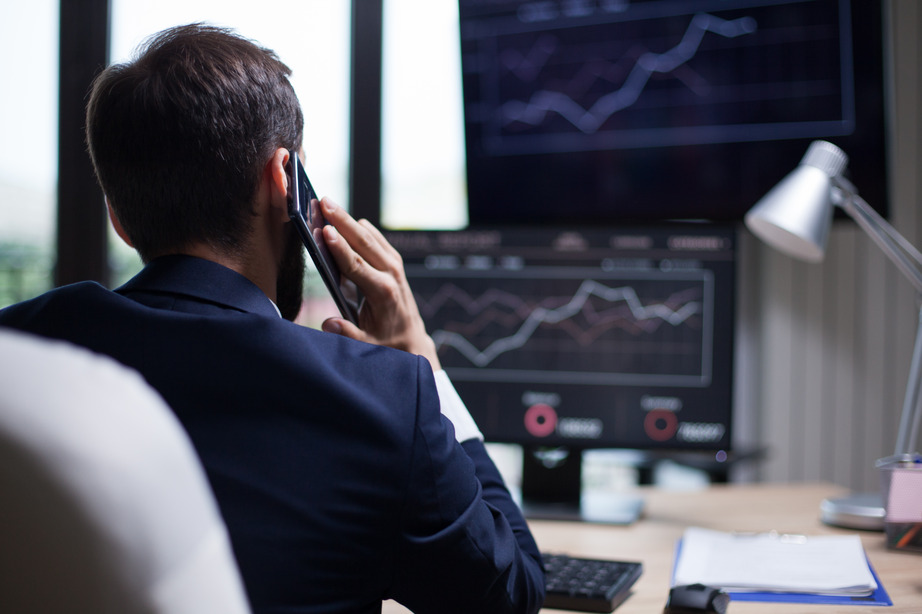 The image shows a man in a suit seated at a desk, viewed from behind. He is on a phone call, with his hand holding a smartphone to his ear. In front of him, several computer monitors display financial charts and data, including line graphs, indicating market trends. The workspace is well-organized, with a lamp on the desk, a keyboard, and some paperwork, suggesting that the person is likely a financial broker or trader monitoring market fluctuations while communicating with a client or colleague. The overall environment conveys a professional, financial setting.