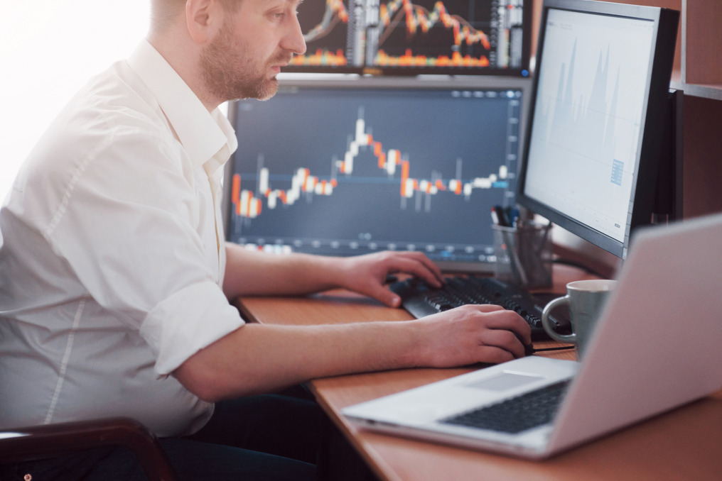 The image shows a man in a white shirt sitting at a desk, focusing intently on his computer screens. He is working with data related to financial markets, as indicated by the stock charts visible on multiple monitors in front of him. His right hand is on the keyboard, and there is a cup next to him on the desk. A laptop is also present, partially visible in the foreground. The setting suggests a work environment related to trading, finance, or data analysis.