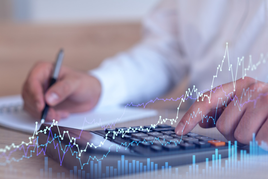 The image shows a person using a calculator, while writing notes in the background. Overlaid on the image is a holographic representation of fluctuating financial data, such as stock or forex charts with various lines and peaks, representing changes in market values. This image conveys the concept of financial analysis or market prediction, with a focus on analyzing historical data to anticipate future trends in trading or investment markets.