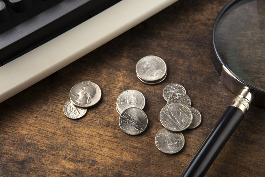 Several U.S. quarters and nickels placed on a wooden surface, with a vintage computer keyboard partially visible on the left and a magnifying glass on the right. The arrangement suggests close examination or study of currency, possibly related to financial analysis or economic research.