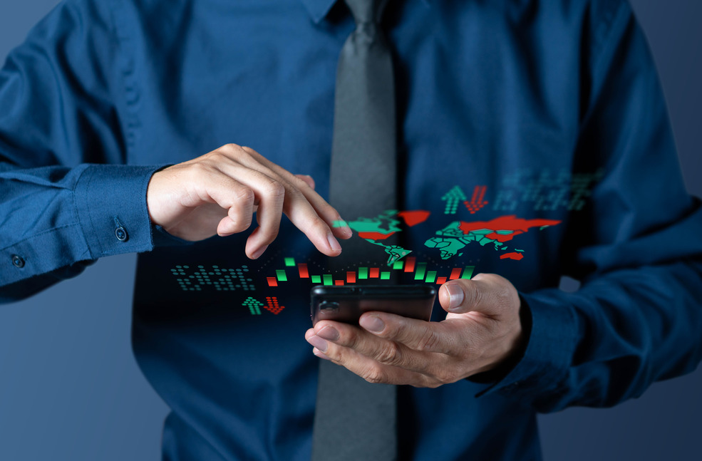 A person in a dark blue shirt and tie is holding a smartphone and interacting with it. Floating above the phone is a holographic display showing a world map with red and green candlestick charts, along with up and down arrows symbolizing stock market trends. The image suggests real-time analysis or monitoring of global stock markets and financial data.