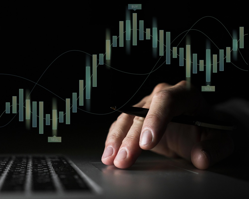 This image showcases a trading or financial concept. It features candlestick charts commonly used in stock market or cryptocurrency analysis. A hand interacts with a laptop, symbolizing data analysis or online trading, with "buy" and "sell" indicators highlighted. The dark background enhances the focus on the chart and the act of trading.