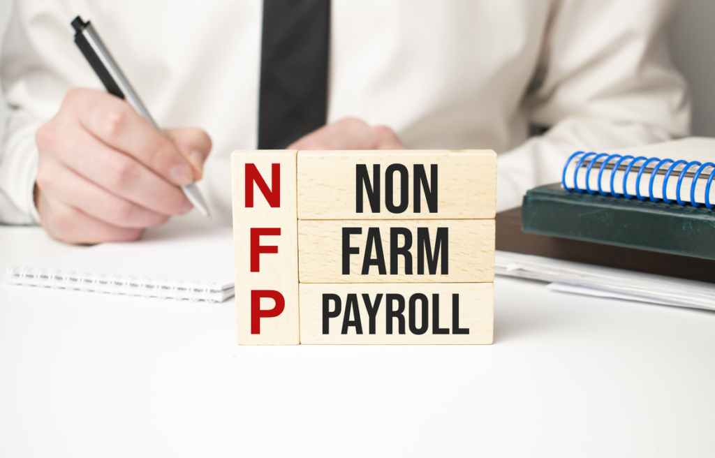 The image shows wooden blocks arranged vertically to spell out "NFP" in red letters on the left and horizontally as "NON FARM PAYROLL" in black letters on the right. In the background, there is a person writing on a notepad, with a blue spiral-bound notebook and additional documents on the desk.