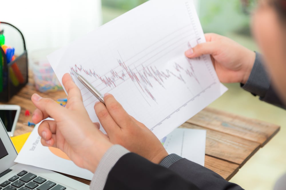 A person in a suit holds and points to a printed financial chart with a pen. The chart shows fluctuating lines, possibly indicating market trends. A laptop and office supplies are visible on the wooden desk.