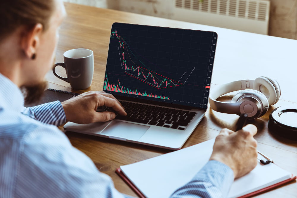 A person in a blue striped shirt is working on a laptop displaying a stock chart with an upward trend line. They hold a pen and have a notebook nearby. Headphones and a cup are on the wooden table.