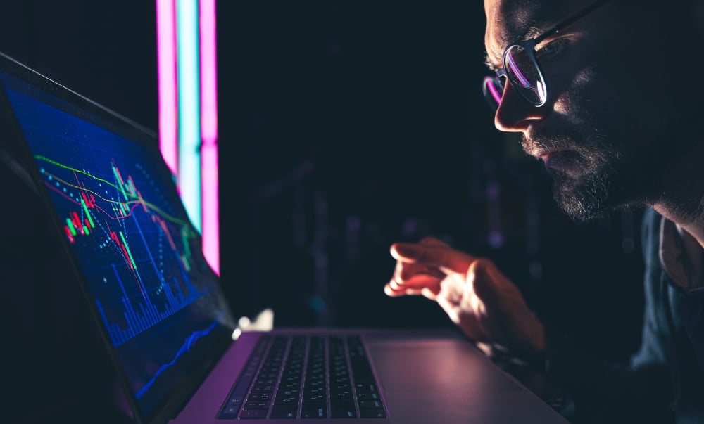 A person wearing glasses closely examines a laptop screen displaying colorful financial charts and graphs in a dimly lit setting. The focus is on the screens data, suggesting analysis or trading activities.