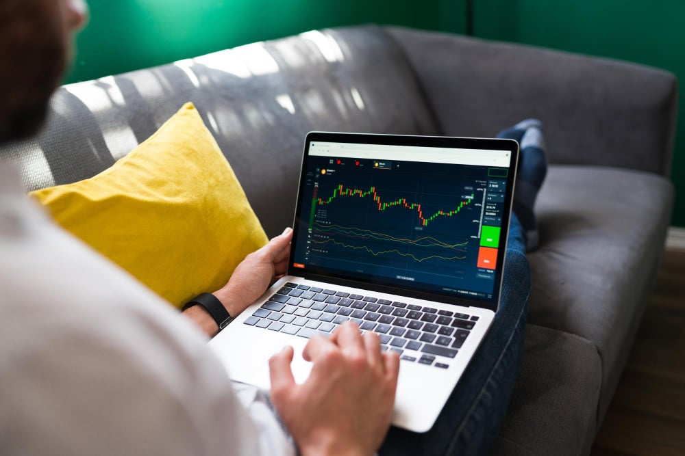 Person sitting on a gray couch with a yellow pillow, using a laptop displaying a financial chart with green and red indicators. The focus is on the screen, suggesting stock market analysis or trading activity.