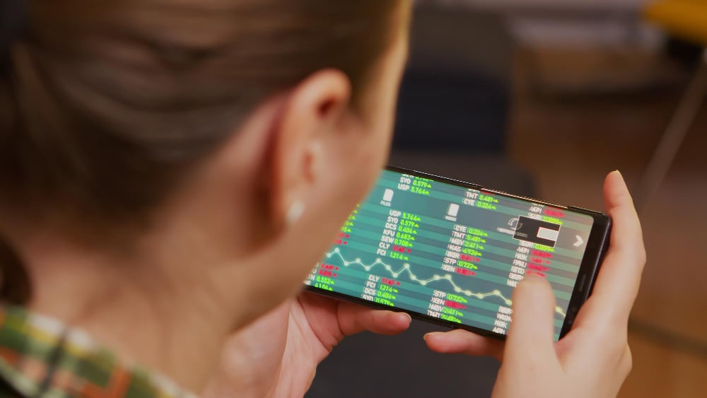 A person holds a smartphone in landscape mode, displaying a screen with a stock market application. The display shows various stock prices and a line graph, indicating financial data trends. The person appears to be closely observing the screen.