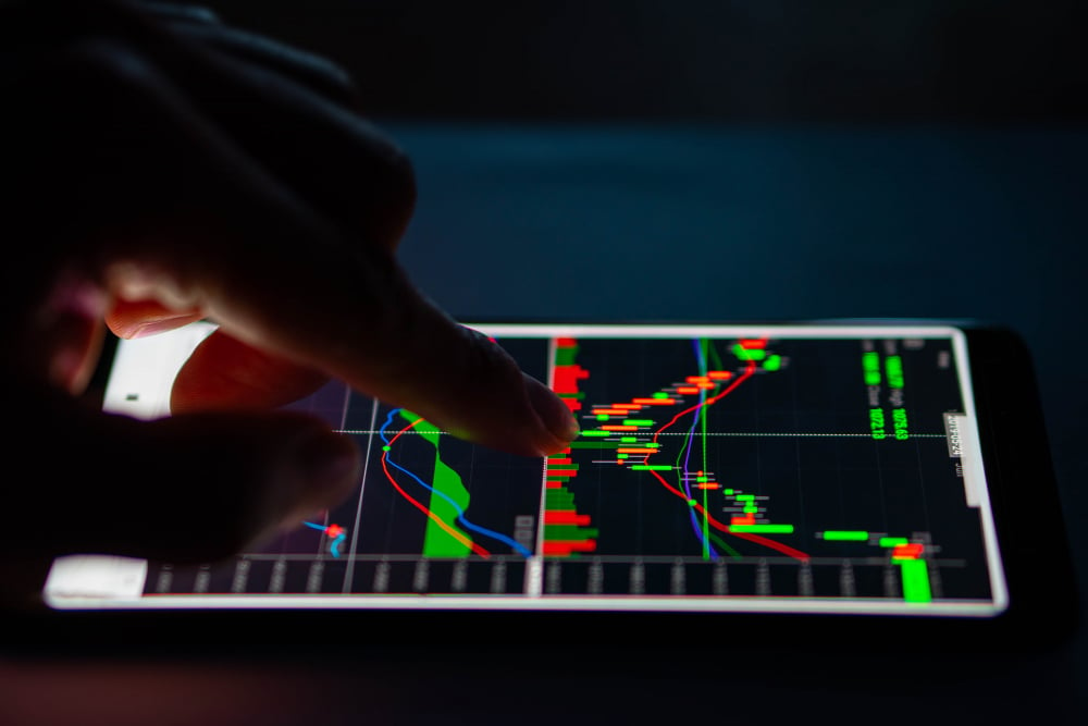 A person interacts with a digital tablet displaying a stock market chart with candlestick patterns and various colored trend lines, indicating financial market data analysis in a dimly lit environment.