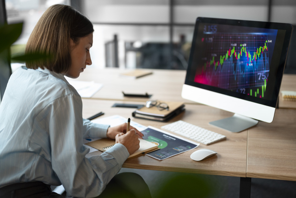 A woman sitting at a desk in an office, analyzing forex charts on a computer screen showing a candlestick chart while taking notes in a notebook.