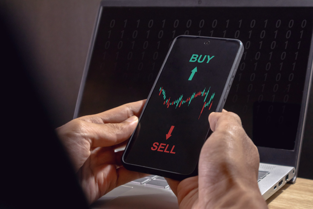 A person holds a phone displaying a stock trading app with "BUY" and "SELL" options and a stock graph, sitting in front of a laptop with binary code on the screen.